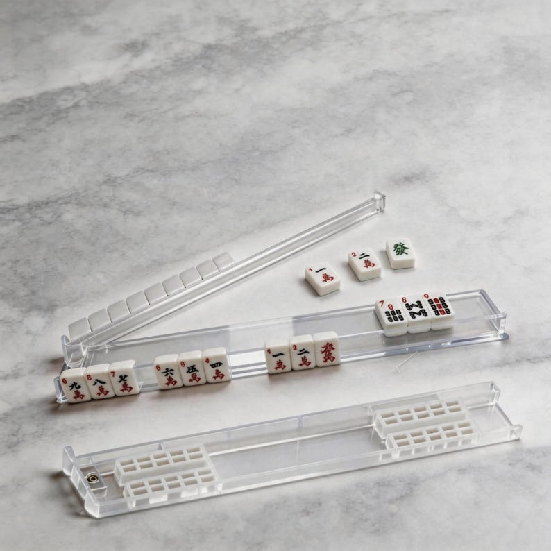 Puede incluir: Un juego de Mahjong de pl&aacute;stico transparente con fichas blancas con s&iacute;mbolos y caracteres rojos y negros. El juego incluye un estante y fichas, dispuestos sobre una superficie de m&aacute;rmol gris claro. El juego es un juego de fichas tradicional.
