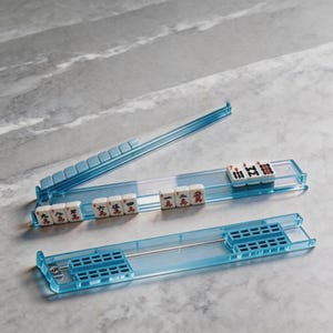 Könnte beinhalten: Ein Set aus hellblauen, transparenten Mahjong-Steinehaltern. Die Halterungen enthalten weiße Spielsteine mit schwarzen und roten Symbolen. Das Set wird auf einer grauen Marmoroberfläche präsentiert. Ideal für Mahjong-Spiele.
