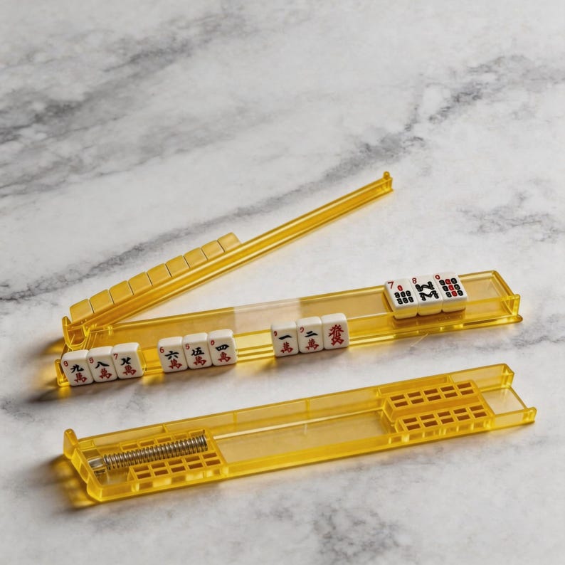 Puede incluir: Un juego de soportes de fichas de Mahjong de pl&aacute;stico amarillo con fichas blancas con caracteres y n&uacute;meros chinos negros. Los soportes son rectangulares con un dise&ntilde;o claro y transparente, y un resorte de metal es visible en uno.