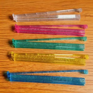 Puede incluir: Una colecci&oacute;n de coloridos clips de pl&aacute;stico para bolsas en transparente, rosa, verde, amarillo y azul. Los clips son rectangulares con un dise&ntilde;o de bisagra para sellar bolsas. Est&aacute;n dispuestos sobre una superficie de madera.