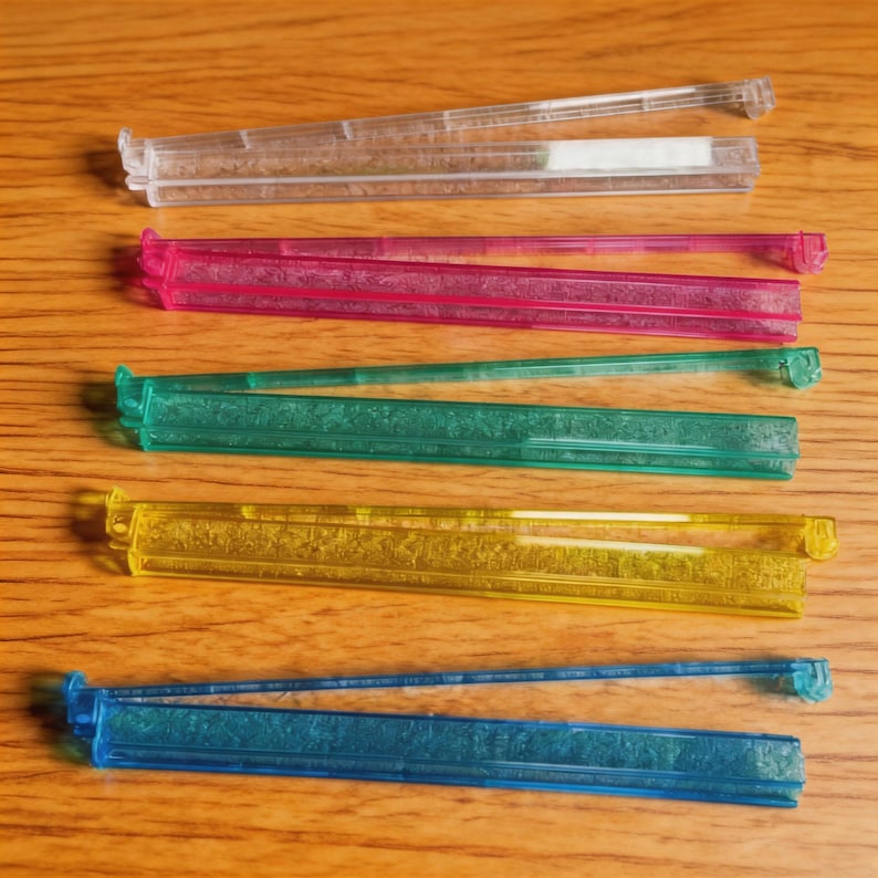 Puede incluir: Una colecci&oacute;n de coloridos clips de pl&aacute;stico para bolsas en transparente, rosa, verde, amarillo y azul. Los clips son rectangulares con un dise&ntilde;o de bisagra para sellar bolsas. Est&aacute;n dispuestos sobre una superficie de madera.