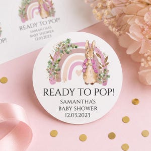 May include: Circular sticker with the text "READY TO POP!" and "SAMANTHA'S BABY SHOWER 12.03.2023". The design includes a rabbit in a pink jacket, a rainbow, and floral elements. The background is a soft pink colour.