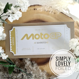 May include: A gold foil ticket with the text "Surprise" and "Happy 50th Birthday Stuart" on the left side. The right side of the ticket says "We are off to MotoGP at Silverstone" with the text "MotoGP" in gold foil. The ticket also says "Staying at the Ridings Campsite" and "23rd - 26th May". The ticket is on a brown background.