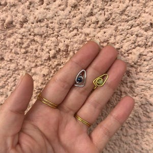 Può includere: Una mano mostra due piccoli orecchini avvolti in filo metallico, uno argentato con una pietra blu scuro e uno dorato con una pietra verde chiaro. Due anelli d'oro adornano le dita. Lo sfondo è una superficie rosa chiaro strutturata.