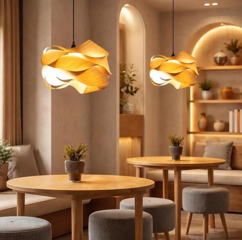 May include: Two pendant lights with a layered, petal-like design hang above a cafe setting. The lights are made of light-colored wood, emitting a warm glow. The cafe features wooden tables, gray stools, and potted plants, creating a cozy atmosphere.