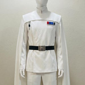 May include: A white costume with a high collar, cape, and black belt. The outfit includes a jacket with a small pocket and a row of coloured pins. The trousers are white, and the costume is displayed on a mannequin.
