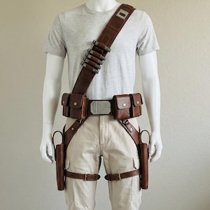 May include: A light gray t-shirt and beige cargo pants worn with a brown leather utility belt system. The gear includes a shoulder strap with metallic cylinders, a central buckle, two waist pouches, and two drop-leg holsters with leg straps.