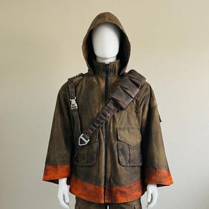 K&ouml;nnte beinhalten: Eine braune Kapuzenjacke mit Used-Optik, einem leuchtend orangefarbenen Streifen an Saum und &Auml;rmelb&uuml;ndchen, einem Frontrei&szlig;verschluss und einem dunkelbraunen Utility-Gurt &uuml;ber der Brust. Das Outfit umfasst eine passende braune Cargohose mit Seitentaschen.