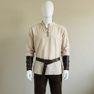 Puede incluir: Una camisa beige de manga larga con cuello acordonado, pantalones de color marr&oacute;n oscuro, un cintur&oacute;n de cuero marr&oacute;n oscuro y brazaletes de antebrazo de cuero marr&oacute;n oscuro a juego con hebillas, sobre un maniqu&iacute; blanco.