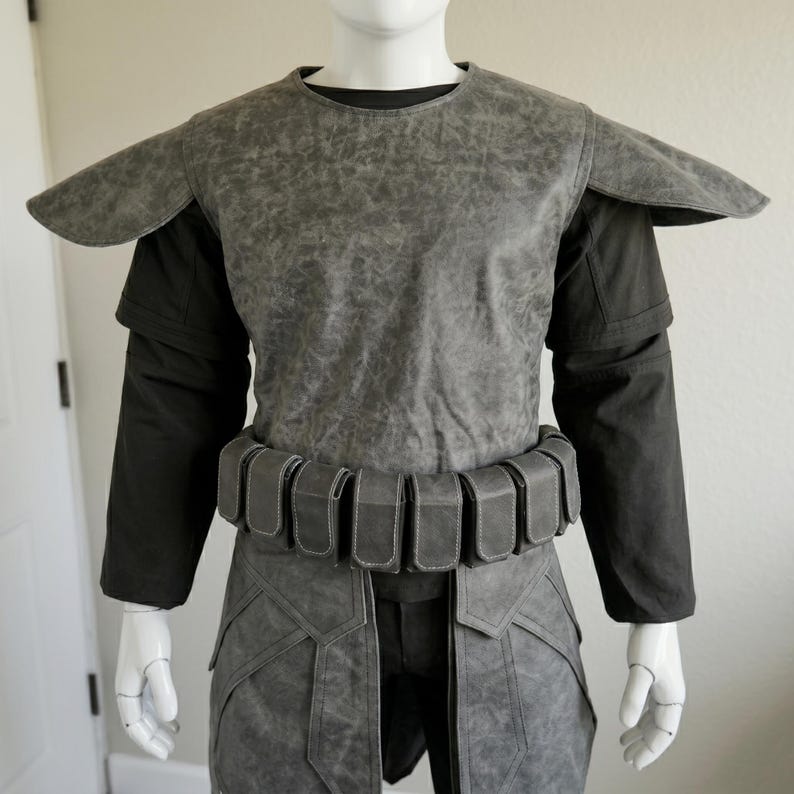 May include: A gray costume with a textured appearance, featuring a top with shoulder pads, long black sleeves, and a belt with multiple pouches. The costume includes a matching gray lower garment.