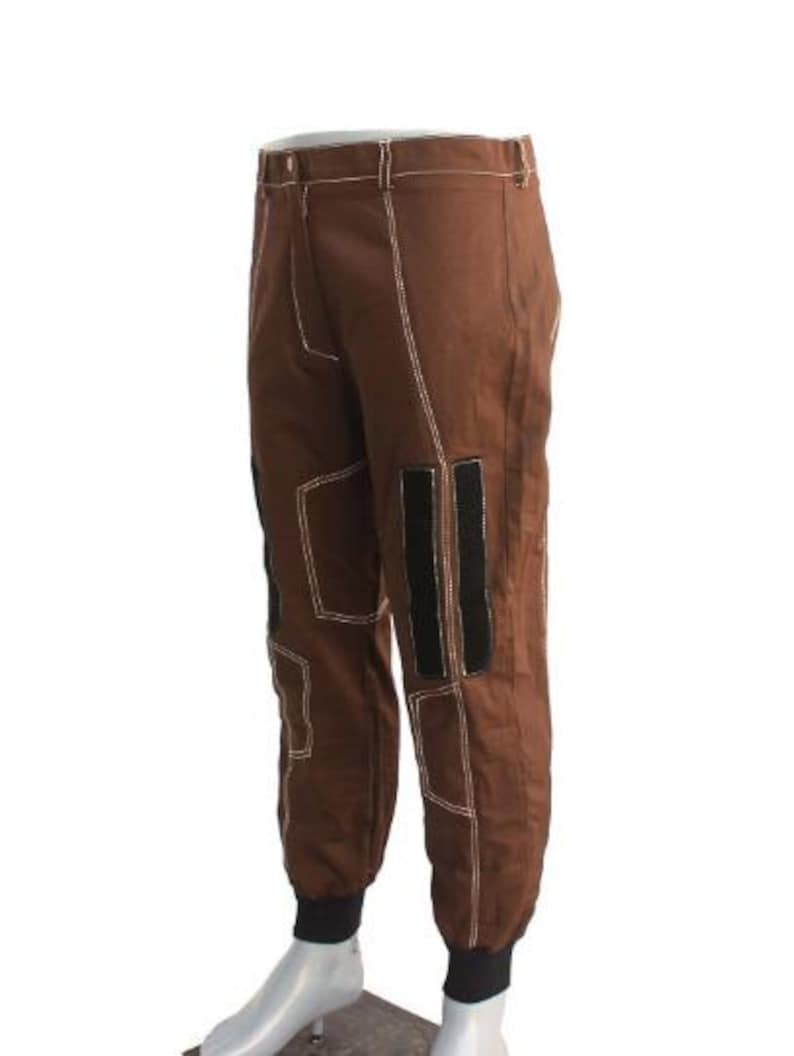 May include: Brown pants with white stitching and black accents. The pants have a classic design with a button and zipper closure, and feature reinforced patches and black stripes on the legs. The cuffs are black.