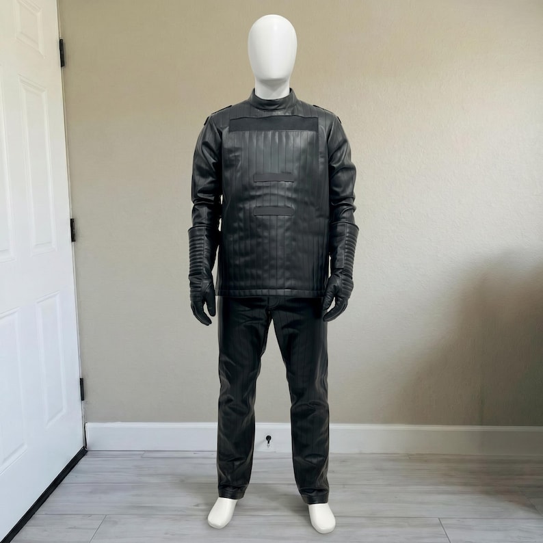 May include: A black leather costume displayed on a mannequin. The outfit includes a long-sleeved jacket with vertical paneling, matching pants, gloves, and white shoes. The jacket has a high collar and ribbed detailing on the sleeves.