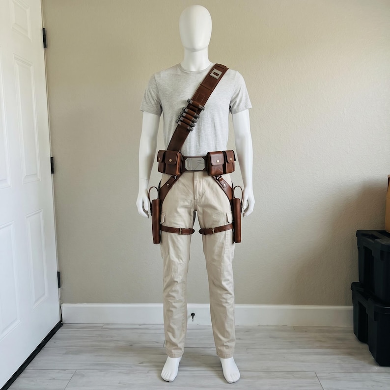 May include: A light gray t-shirt and beige cargo pants worn with a brown leather utility belt system. The gear includes a shoulder strap with metallic cylinders, a central buckle, two waist pouches, and two drop-leg holsters with leg straps.
