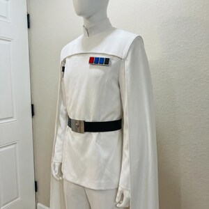 Puede incluir: Traje blanco con cuello alto, mangas tipo capa y cintur&oacute;n negro con hebilla plateada. El uniforme presenta una serie de insignias rectangulares de colores en el pecho. El disfraz se exhibe en un maniqu&iacute;.