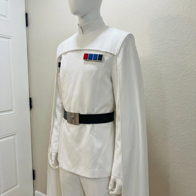 Puede incluir: Traje blanco con cuello alto, mangas tipo capa y cintur&oacute;n negro con hebilla plateada. El uniforme presenta una serie de insignias rectangulares de colores en el pecho. El disfraz se exhibe en un maniqu&iacute;.