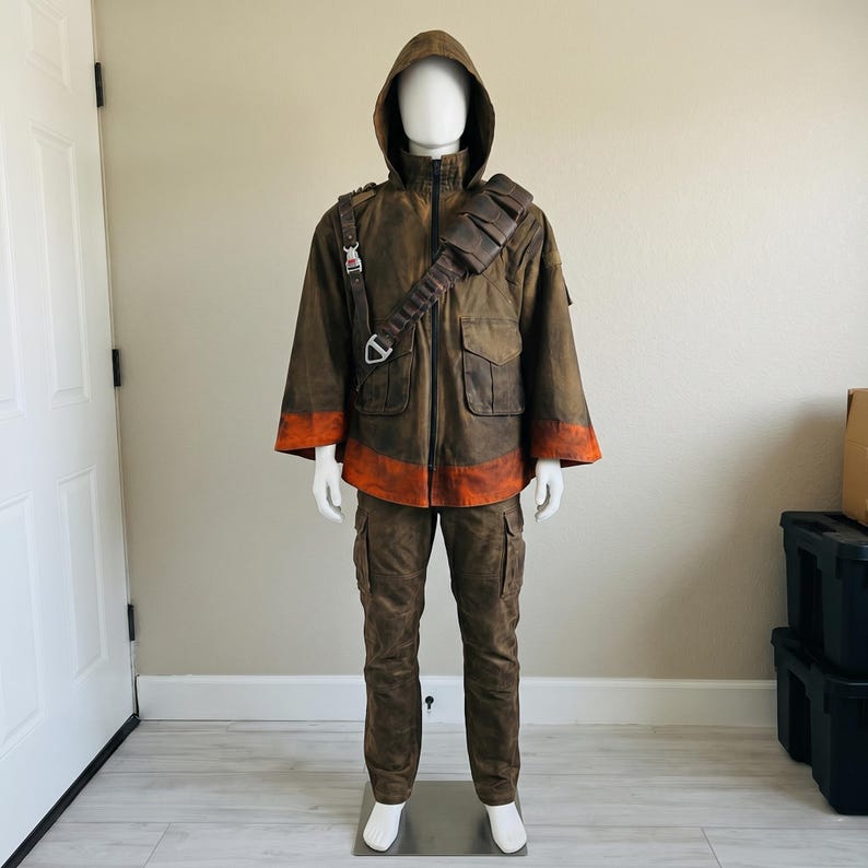 K&ouml;nnte beinhalten: Eine braune Kapuzenjacke mit Used-Optik, einem leuchtend orangefarbenen Streifen an Saum und &Auml;rmelb&uuml;ndchen, einem Frontrei&szlig;verschluss und einem dunkelbraunen Utility-Gurt &uuml;ber der Brust. Das Outfit umfasst eine passende braune Cargohose mit Seitentaschen.
