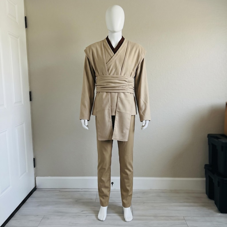 May include: A tan Jedi costume on a mannequin. The outfit features a long-sleeved tunic with a wrap-around belt, matching trousers, and a dark brown undershirt. The costume is designed to resemble a Jedi Knight's attire, suitable for cosplay.