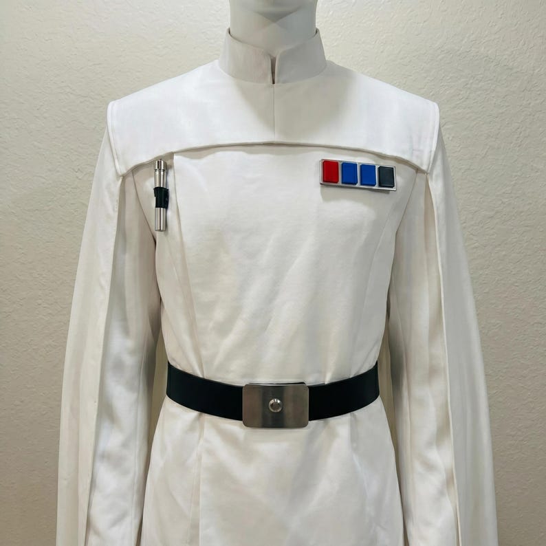 Puede incluir: Un uniforme blanco con cuello mandar&iacute;n, capa en los hombros y cintur&oacute;n negro. El uniforme presenta un portal&aacute;pices plateado y una insignia rectangular con cuadrados rojos, azules y grises. El cintur&oacute;n tiene una hebilla plateada.