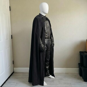 May include: A black costume displayed on a mannequin. The costume includes a long cape, a leather-like chest piece, pants, gloves, and a belt. The mannequin is wearing white shoes. The costume is designed for cosplay or costume parties.