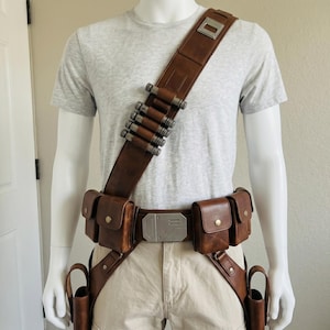 May include: A brown leather utility belt and bandolier set featuring multiple pouches, a silver-toned rectangular buckle, and a diagonal strap across the chest holding five metallic cylindrical canisters. The gear is worn over a light gray t-shirt.