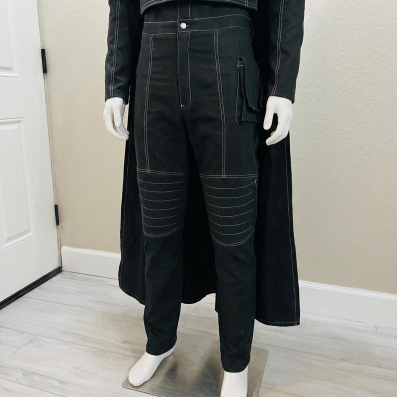 May include: Black pants with white stitching and a matching jacket are displayed on a mannequin. The pants have a pocket on the right leg and horizontal stitching on the knees. The jacket has white stitching and a button closure. A black cape is draped behind the outfit.