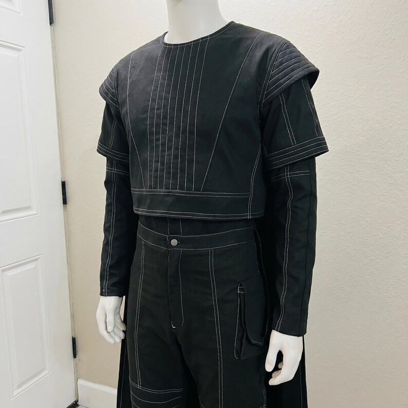 May include: Black two-piece outfit with white stitching. The top is a cropped design with long sleeves and a round neckline. The pants are high-waisted with a button closure and pockets. The outfit is displayed on a mannequin.