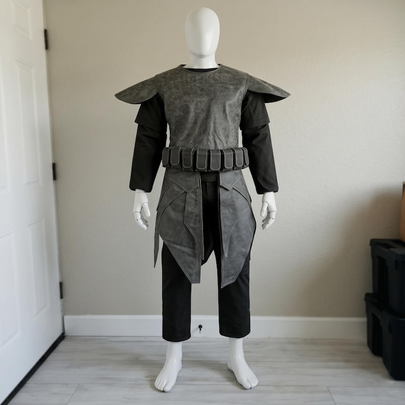May include: A grey and black costume on a mannequin. The outfit features a structured grey top with shoulder pads, a belt with pouches, and a layered skirt. The mannequin also wears a black long-sleeved shirt and black trousers.