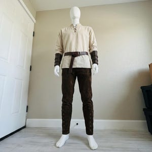 Puede incluir: Una t&uacute;nica beige de manga larga con cuello acordonado, pantalones de color marr&oacute;n oscuro con botones laterales, un cintur&oacute;n de cuero marr&oacute;n oscuro y brazaletes de cuero marr&oacute;n oscuro a juego con hebillas sobre un maniqu&iacute; blanco.