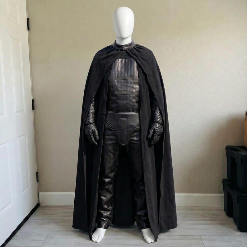 May include: A black costume comprising a long cape, gloves, and trousers, displayed on a mannequin. The outfit has a leather-like appearance, a high collar, and a belt. The gloves have a textured design. The costume is all black.