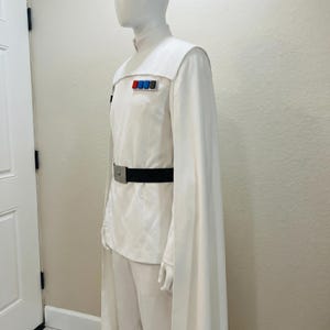 Puede incluir: Un disfraz blanco con cuello alto, mangas tipo capa y cintur&oacute;n negro. El uniforme presenta una serie de insignias rectangulares de colores en el pecho. El disfraz se exhibe en un maniqu&iacute;.