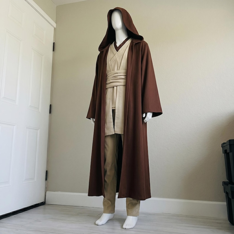 May include: A brown hooded robe with a long, flowing design, paired with a tan tunic, trousers, and a wide sash. The costume is displayed on a mannequin, suggesting a cosplay or costume theme.