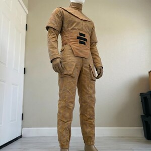 K&ouml;nnte beinhalten: Ein beigefarbenes Kost&uuml;m mit hohem Kragen, Schulterakzenten und einem breiten G&uuml;rtel mit drei schwarzen Rechtecken. Das Kost&uuml;m umfasst lange &Auml;rmel, Handschuhe und eine Hose mit Taschen. Das Outfit wird auf einer Schaufensterpuppe pr&auml;sentiert.