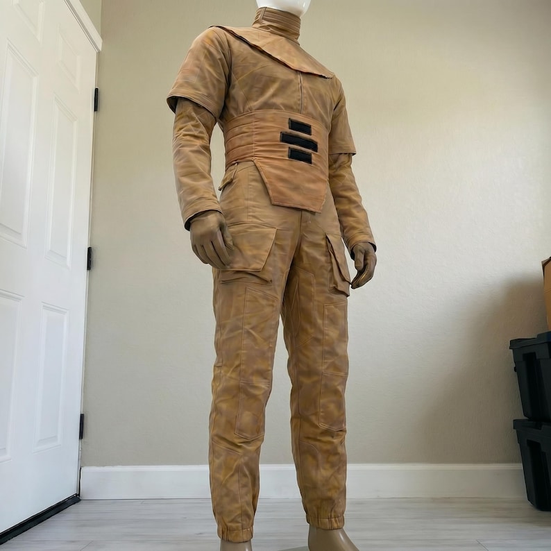 K&ouml;nnte beinhalten: Ein beigefarbenes Kost&uuml;m mit hohem Kragen, Schulterakzenten und einem breiten G&uuml;rtel mit drei schwarzen Rechtecken. Das Kost&uuml;m umfasst lange &Auml;rmel, Handschuhe und eine Hose mit Taschen. Das Outfit wird auf einer Schaufensterpuppe pr&auml;sentiert.