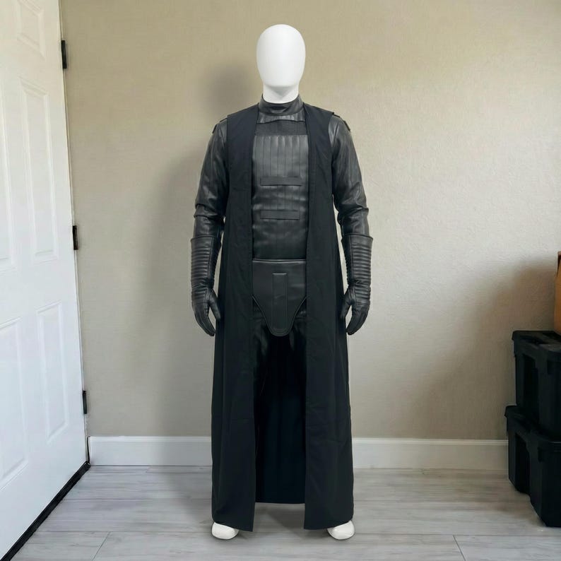 May include: A black costume displayed on a mannequin. The costume includes a long coat, gloves, and a chest piece. The outfit is made of a combination of materials, with a sleek, dark appearance. The mannequin is wearing white shoes.