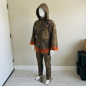 K&ouml;nnte beinhalten: Ein braunes Set aus Kapuzenjacke und Hose mit verwitterter Textur, hohem Kragen und einem diagonalen Brustgurt mit Taschen. Der Saum der Jacke und die &Auml;rmelb&uuml;ndchen sind mit einem markanten orangefarbenen Band akzentuiert.