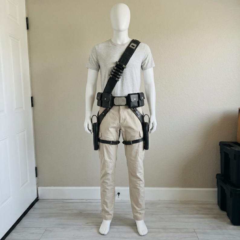 May include: A mannequin wearing a light gray t-shirt and beige cargo pants, equipped with a black tactical utility belt system. The gear includes a shoulder strap with ammunition loops, a central buckle, two waist pouches, and two leg holsters.
