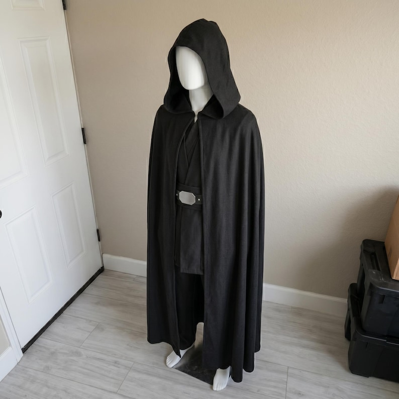 May include: A full-length black hooded cloak worn over a black tunic and trousers, featuring a wide belt with a prominent rectangular silver-toned buckle at the center. The ensemble is displayed on a mannequin, showcasing a dark, layered aesthetic.