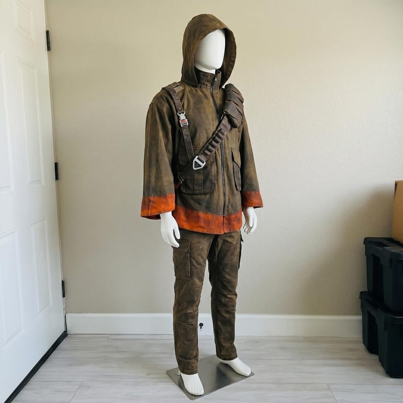 K&ouml;nnte beinhalten: Ein braunes Set aus Kapuzenjacke und Hose mit abgenutzter Textur, hohem Kragen und orangefarbenen B&auml;ndern an &Auml;rmelb&uuml;ndchen und Saum. Die Jacke verf&uuml;gt &uuml;ber einen diagonalen Brustgurt mit mehreren Schlaufen und einen Frontrei&szlig;verschluss.