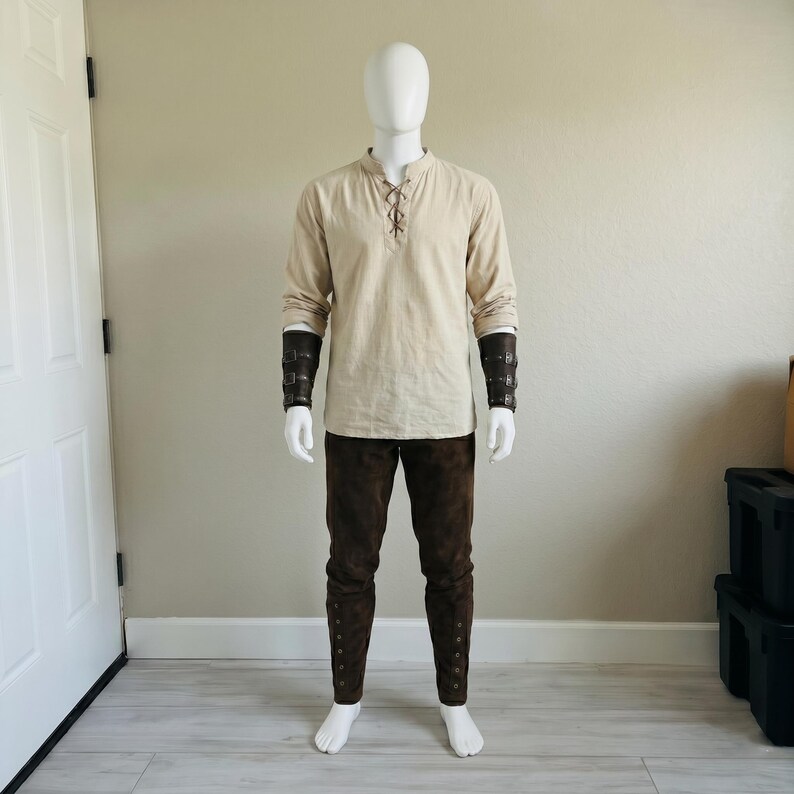 Puede incluir: Una camisa beige de manga larga con cuello acordonado, pantalones de color marr&oacute;n oscuro con botones laterales y brazaletes de antebrazo de cuero sint&eacute;tico marr&oacute;n oscuro con hebillas met&aacute;licas sobre un maniqu&iacute; blanco.