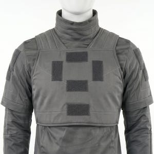 May include: A gray tactical vest with a high collar and multiple rectangular patches. The vest has a padded design and is worn over a long-sleeved garment. The overall aesthetic suggests a military or futuristic theme.