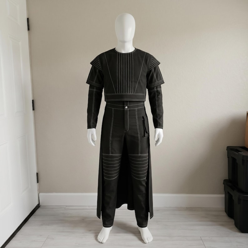 May include: A black two-piece outfit with white stitching details. The top is a cropped jacket with long sleeves and a round neckline. The trousers are high-waisted with a long skirt-like panel at the back. The outfit is displayed on a mannequin.