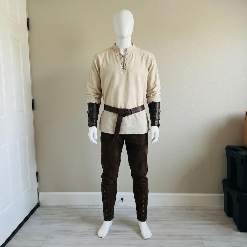 Puede incluir: Una camisa beige de manga larga con cuello acordonado, pantalones de color marr&oacute;n oscuro, un cintur&oacute;n de cuero marr&oacute;n oscuro y brazaletes de antebrazo de cuero marr&oacute;n oscuro a juego con hebillas, sobre un maniqu&iacute; blanco.