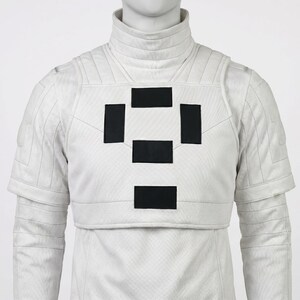 Puede incluir: Chaqueta blanca de estilo futurista con cuello alto y mangas cortas. La chaqueta presenta una serie de parches rectangulares negros en el pecho. La prenda se exhibe en un maniqu&iacute;.