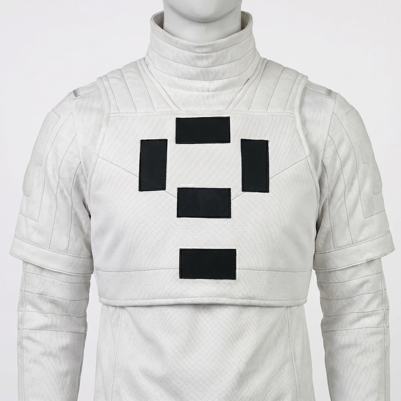 Puede incluir: Chaqueta blanca de estilo futurista con cuello alto y mangas cortas. La chaqueta presenta una serie de parches rectangulares negros en el pecho. La prenda se exhibe en un maniqu&iacute;.