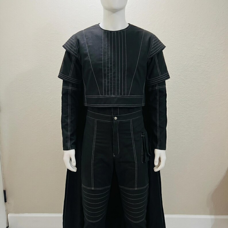 May include: Black two-piece outfit with white stitching detail. The top is a cropped jacket with layered shoulders and long sleeves. The pants are high-waisted with knee paneling. A long black cape is attached to the back of the jacket.