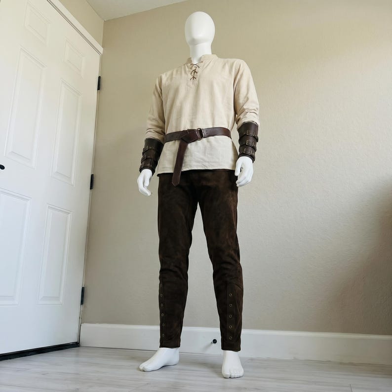 Puede incluir: Una t&uacute;nica beige de manga larga con cuello acordonado, pantalones de color marr&oacute;n oscuro, un cintur&oacute;n de cuero marr&oacute;n con una anilla met&aacute;lica y brazaletes de cuero marr&oacute;n a juego con hebillas, sobre un maniqu&iacute; blanco ante una pared.