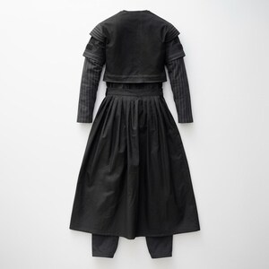 May include: A black outfit comprising a cropped jacket, a long-sleeved shirt with pinstripes, a pleated skirt, and matching trousers. The jacket has a round neckline and layered sleeves. The skirt is long and flowing, and the trousers are straight-legged.