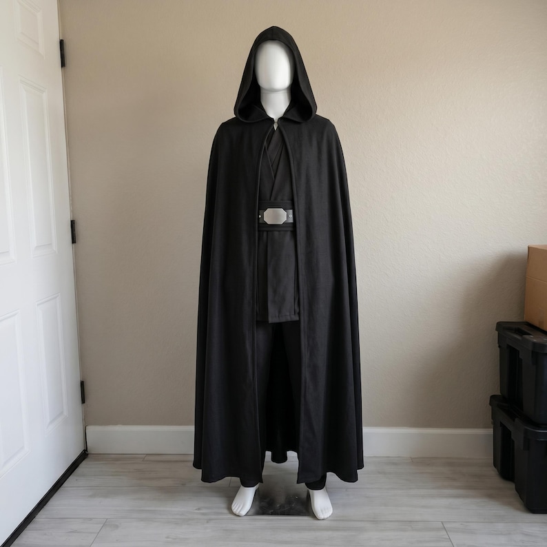 May include: A full-length black hooded cloak worn over a black tunic and trousers, featuring a wide black belt with a rectangular silver-toned metallic buckle at the center, displayed on a white mannequin against a plain interior wall.