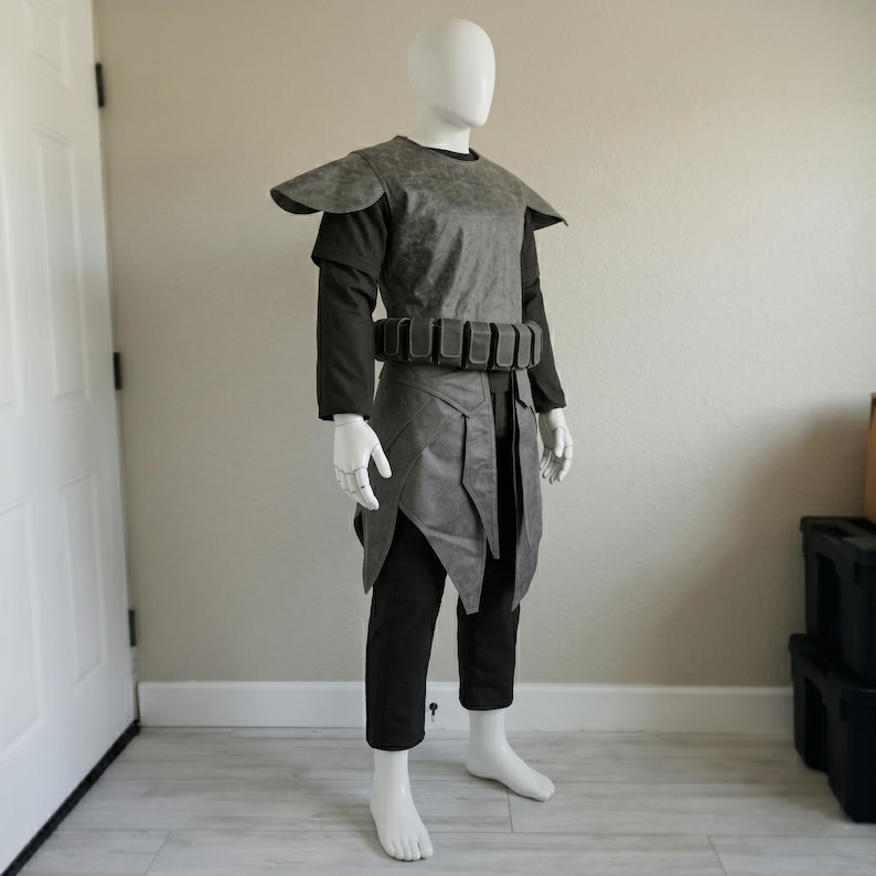 May include: A gray and black costume displayed on a mannequin. The costume includes a gray top with shoulder pads, a belt with pouches, black pants, and a gray skirt-like piece. The mannequin is standing on a light-colored floor.