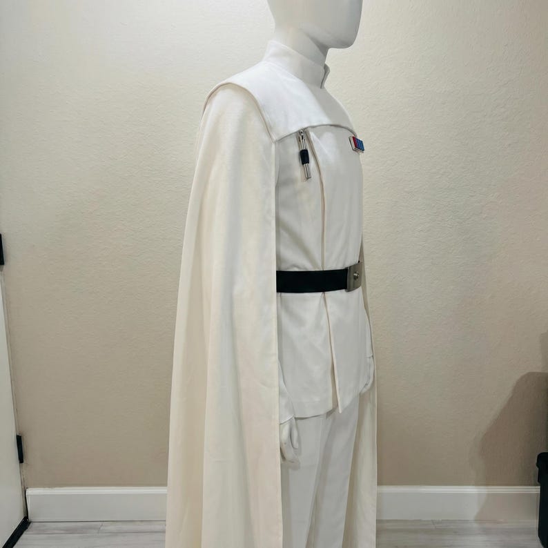 Puede incluir: Traje color crema con capa a juego. El atuendo incluye una chaqueta de cuello alto con cintur&oacute;n negro y hebilla plateada, y pantalones a juego. Se ve una peque&ntilde;a insignia en la chaqueta. El disfraz se exhibe en un maniqu&iacute;.